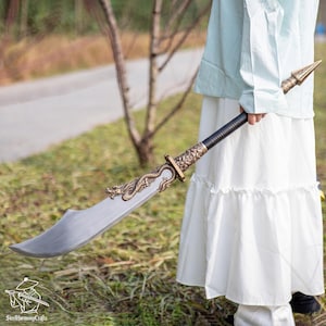 May include: A decorative sword with a curved, silver blade and ornate gold-colored dragon detailing. The handle is black with gold accents. The sword is held by a person wearing a white dress and a light blue top.
