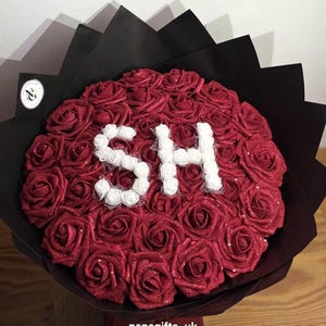 May include: A circular bouquet of deep red glittery artificial roses wrapped in black paper. The centre of the arrangement features the letters SH formed from small white artificial roses. A small circular logo is visible on the left side.
