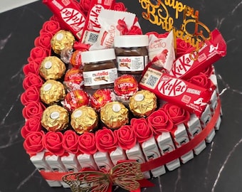 May include: A heart-shaped gift arrangement with red roses, Kit Kat bars, Ferrero Rocher chocolates, Nutella jars, and Raffaello candies. A gold "Happy Birthday" topper and a red ribbon with a bow complete the festive presentation.