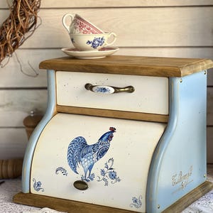 Può includere: Un portapane in legno con cornice azzurra e pannelli color crema. La parte anteriore presenta un'illustrazione di un gallo blu e un pomello in metallo. Include un cassetto superiore con maniglia in ceramica. Due tazze da tè sono sopra.