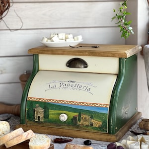 May include: A rectangular wooden bread bin with a cream-coloured roll-top door featuring the text La Panetteria and a painted landscape of a building and trees. The bin has dark green side panels, a natural wood top, and a small white knob.