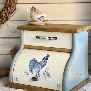 May include: A wooden bread box with a light blue frame and cream-colored panels. The front features a blue rooster illustration and a metal handle. A drawer sits above the roll-top door, which is adorned with a matching blue floral ceramic pull.