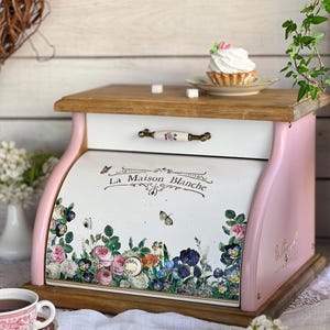 Op de afbeelding: Een roze en witte broodtrommel met een roldeur voorzien van een bloemenprint en de tekst La Maison Blanche. De unit bevat een bovenste lade met een keramische handgreep, een basis van natuurlijk hout en een bijpassend houten bovenvlak.