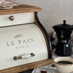 May include: Cream-colored bread box with the text "LE PAIN PARIS" and a decorative handle. The box is made of wood and metal, with a black trim. A cup of coffee, a coffee maker, and a sugar cube are also visible.