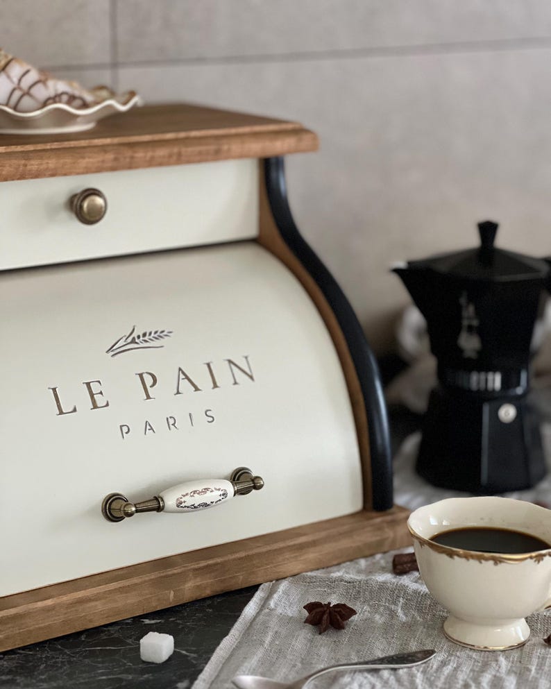 May include: Cream-colored bread box with the text "LE PAIN PARIS" and a decorative handle. The box is made of wood and metal, with a black trim. A cup of coffee, a coffee maker, and a sugar cube are also visible.