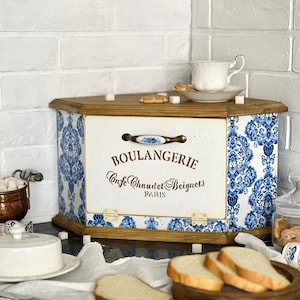 May include: A wooden corner bread bin with a white and blue floral pattern. The bin has a wooden top and base, with the words "BOULANGERIE Cafe Chaudet Beignets PARIS" on the front. A white teacup and saucer are on top.