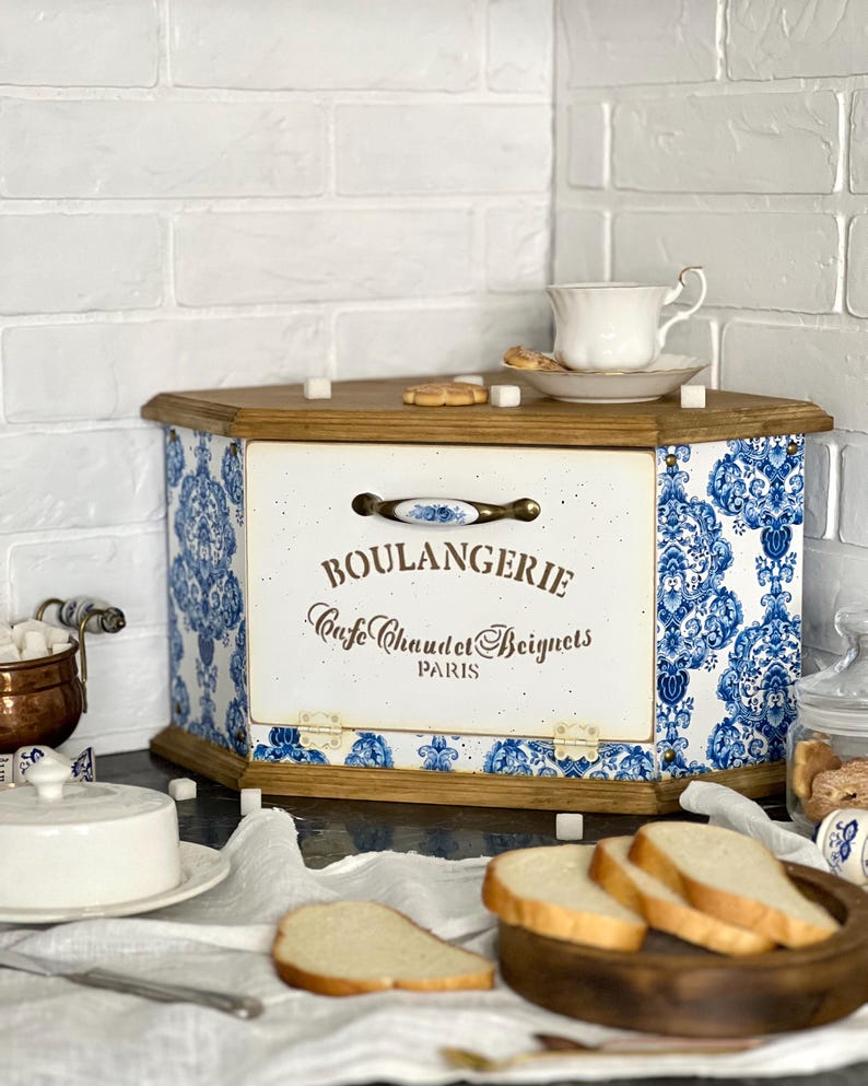 May include: A wooden bread box with a hinged front door featuring blue and white damask patterns. The front panel displays the text BOULANGERIE, Cafe Chaud et Beignets, and PARIS in brown lettering, accented by a decorative ceramic handle and metal hinges.