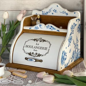 Peut inclure: Un coffre à pain blanc et bois avec des motifs floraux bleus. Le texte "La Boulangerie Paris" est affiché dans un cadre décoratif. Le design comprend un dessus incurvé et une base en bois, avec une poignée à l'avant.