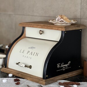 May include: Cream and black bread box with a wooden top and base. The box has a drawer and a curved front with the words "LE PAIN PARIS" printed on it. A small plate with food sits on top. The box has a decorative handle.