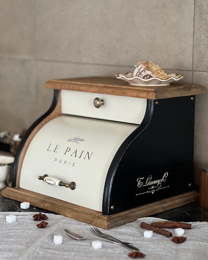 May include: Cream and black bread box with a wooden top and base. The box has a drawer and a curved front with the words "LE PAIN PARIS" printed on it. A small plate with food sits on top. The box has a decorative handle.