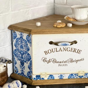 May include: A wooden bread bin with a white front panel featuring the text BOULANGERIE, Cafe Chaud et Beignets, PARIS. The bin has blue and white damask patterned sides and base, a metal handle with a ceramic insert, and a natural wood top.
