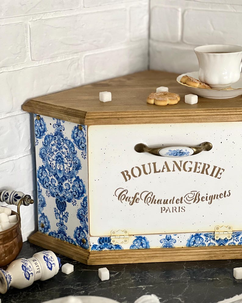 May include: A wooden bread bin with a white front panel featuring the text BOULANGERIE, Cafe Chaud et Beignets, PARIS. The bin has blue and white damask patterned sides and base, a metal handle with a ceramic insert, and a natural wood top.