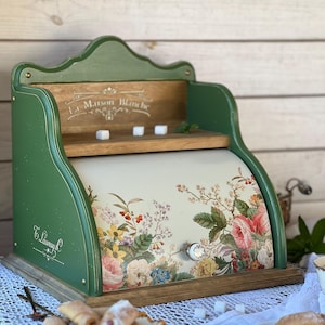 May include: A green wooden bread box with a curved, cream-colored roll-top door featuring a floral print of roses and wildflowers. The side is inscribed with the text 'La Maison Blanche' and 'T. Launay'. It includes a small shelf and a round knob.