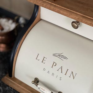 May include: Cream-colored bread box with the text "LE PAIN PARIS" and a wheat sprig design. The box has a wooden top and base, a drawer with a brass knob, and a decorative handle. A dried red chili pepper rests on the top.