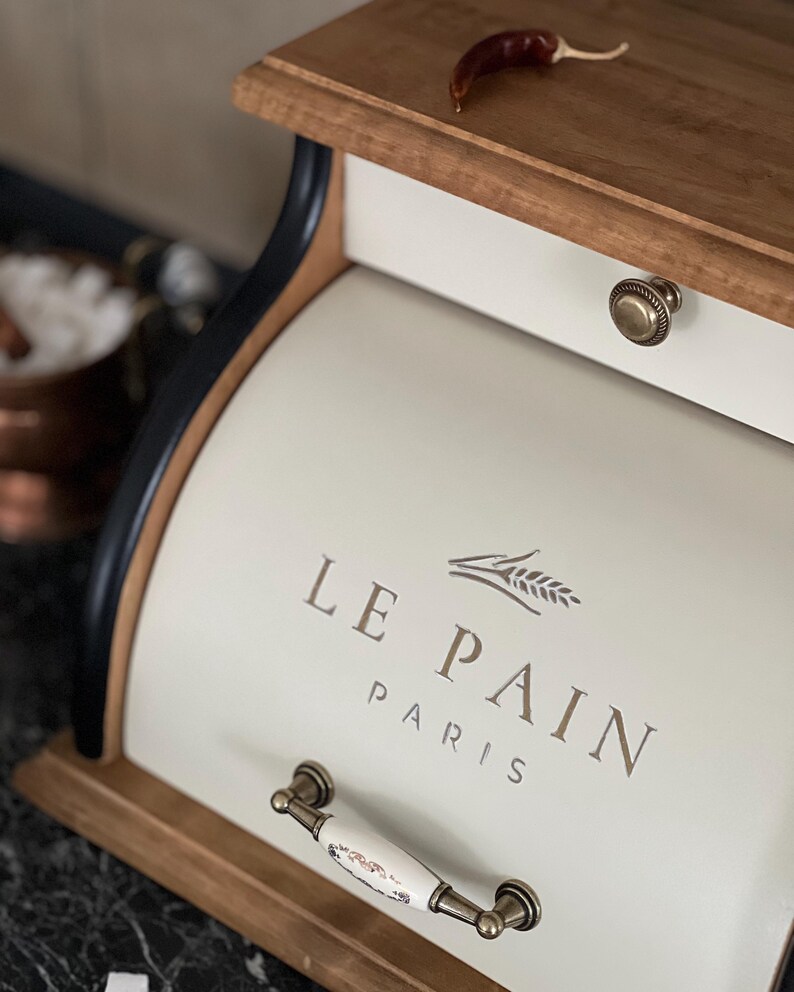 May include: Cream-colored bread box with the text "LE PAIN PARIS" and a wheat sprig design. The box has a wooden top and base, a drawer with a brass knob, and a decorative handle. A dried red chili pepper rests on the top.