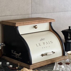 May include: A wooden bread box with a cream-colored front and a black curved side. The words "LE PAIN PARIS" are printed on the front. The box has a wooden top and base, and a small drawer. A decorative handle is on the front.