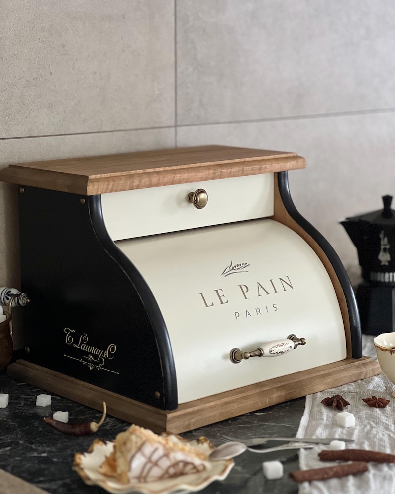 May include: A wooden bread box with a cream-colored front and a black curved side. The words "LE PAIN PARIS" are printed on the front. The box has a wooden top and base, and a small drawer. A decorative handle is on the front.