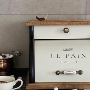 May include: Cream-colored bread box with the text "LE PAIN PARIS" in brown lettering. The box has a wooden top and base, black side accents, and a decorative handle. A small red pepper sits on top.