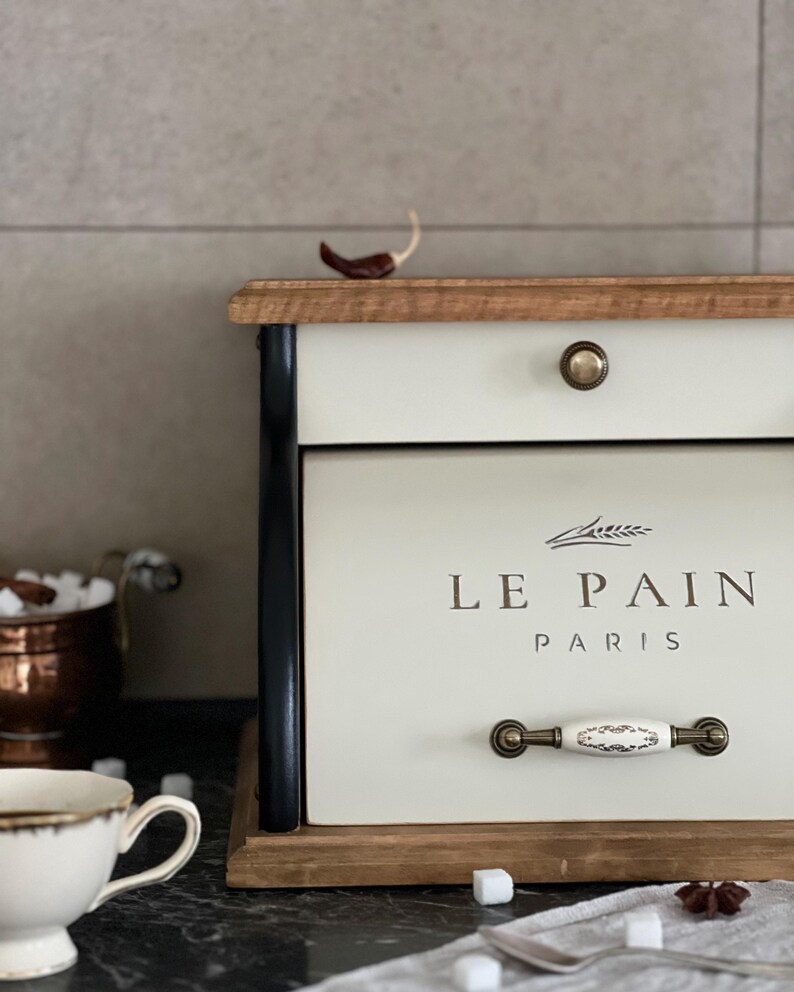 May include: Cream-colored bread box with the text "LE PAIN PARIS" in brown lettering. The box has a wooden top and base, black side accents, and a decorative handle. A small red pepper sits on top.