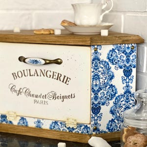 May include: A wooden bread box with a cream-coloured front panel featuring the text BOULANGERIE Cafe Chaud et Beignets PARIS. The sides are decorated with a blue and white floral pattern, and it includes a metal handle with a floral accent.