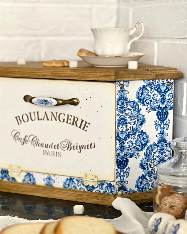 May include: A wooden bread box with a cream-coloured front panel featuring the text BOULANGERIE Cafe Chaud et Beignets PARIS. The sides are decorated with a blue and white floral pattern, and it includes a metal handle with a floral accent.