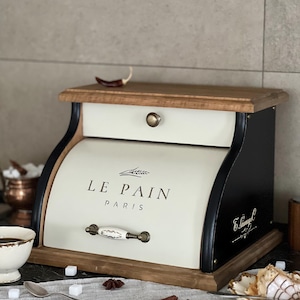 May include: Cream-colored bread box with a wooden top and base, and black curved sides. The front of the box features the text "LE PAIN PARIS" in black. A decorative handle and knob are visible.