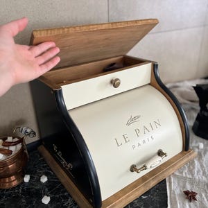 May include: Cream-colored bread box with a wooden lid and base, and black trim. The text "LE PAIN PARIS" is printed on the front. The box has a drawer and a handle with a white ceramic finish.