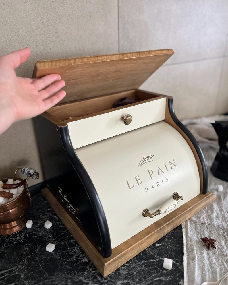 May include: Cream-colored bread box with a wooden lid and base, and black trim. The text "LE PAIN PARIS" is printed on the front. The box has a drawer and a handle with a white ceramic finish.
