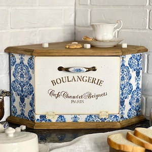May include: A white and blue corner bread bin with a wooden top and base. The bin features a blue floral pattern and the words "BOULANGERIE Cafe Chaudet Beignets PARIS". A teacup and saucer sit on top.