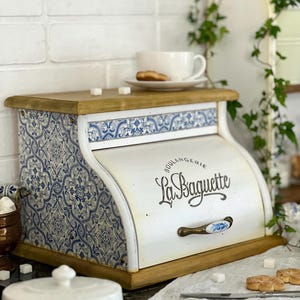 May include: A wooden bread bin with a roll-top door featuring the text "Boulangerie La Baguette" in black script. The sides are decorated with a blue and white floral pattern, and the front handle is white with a blue floral design.