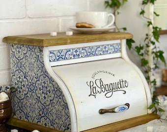 May include: A wooden bread box with a roll-top door featuring the text "Boulangerie La Baguette" in black script. The sides are decorated with a blue and white floral pattern, and the front handle is white with a blue floral design.