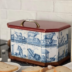 May include: A rectangular bread bin with a red lid and a wooden base. The sides are decorated with blue and white tiles depicting pastoral scenes. A gold-coloured handle is attached to the top.