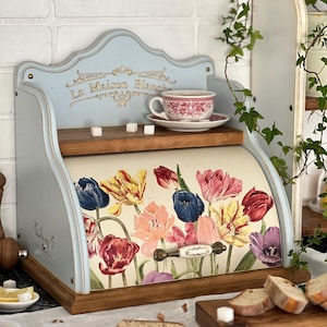 May include: A vintage-style bread bin in light blue, featuring a floral tulip design. It has a wooden base and a handle. A teacup and saucer are on a shelf above. The text "La Maison Blanche" is visible.
