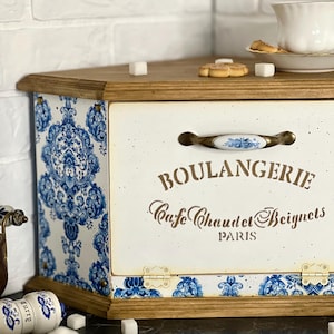 May include: A wooden bread box with a cream-coloured front panel featuring the text BOULANGERIE Cafe Chaud et Beignets PARIS. The sides and bottom edge are decorated with a blue and white damask pattern. It has a central handle with blue floral accents.