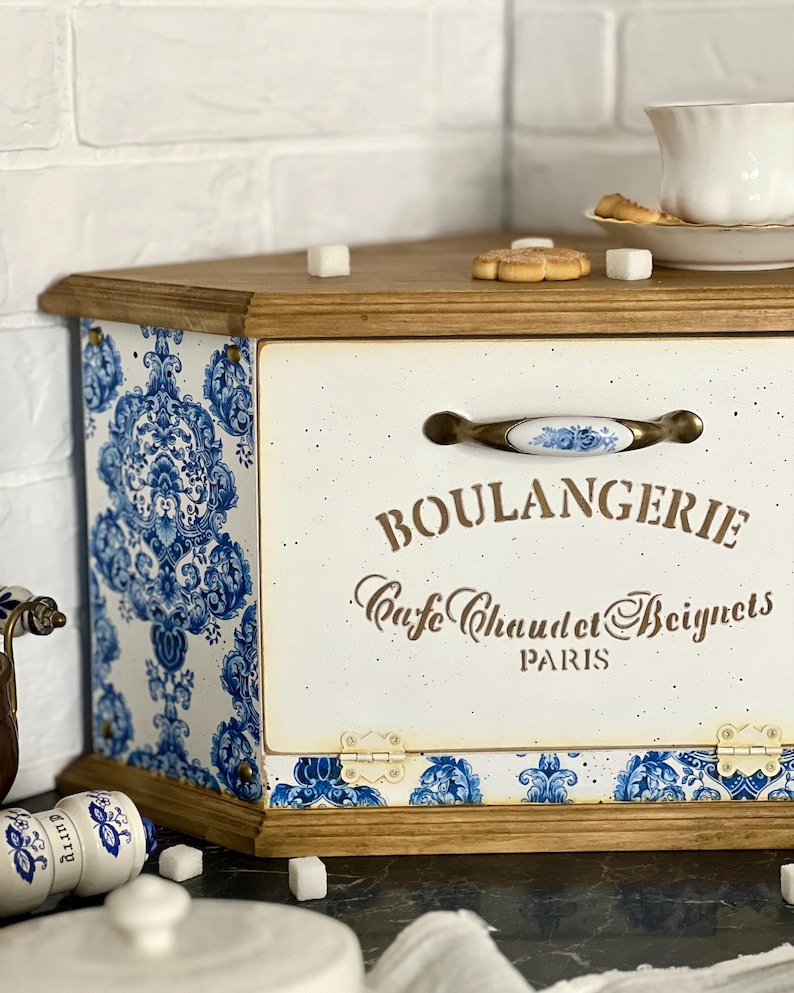 May include: A wooden bread box with a cream-coloured front panel featuring the text BOULANGERIE Cafe Chaud et Beignets PARIS. The sides and bottom edge are decorated with a blue and white damask pattern. It has a central handle with blue floral accents.