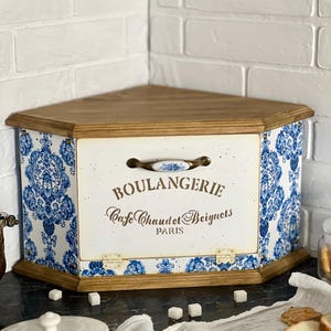 May include: A corner bread bin with a wooden top and base, featuring a white front panel with blue and white damask patterned sides. The front displays the text BOULANGERIE, Cafe Chaud et Beignets, PARIS, and includes a ceramic handle with blue floral accents.