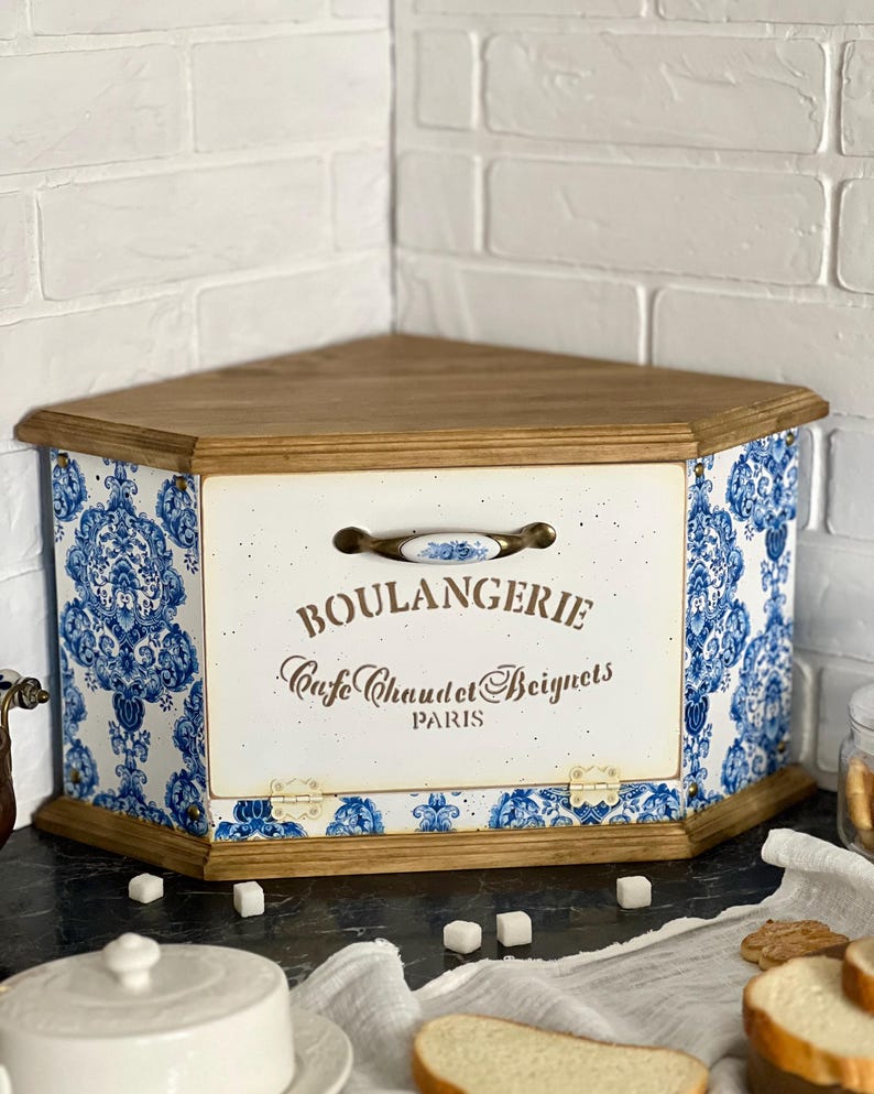 May include: A corner bread bin with a wooden top and base, featuring a white front panel with blue and white damask patterned sides. The front displays the text BOULANGERIE, Cafe Chaud et Beignets, PARIS, and includes a ceramic handle with blue floral accents.