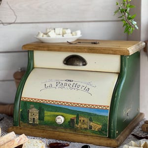 May include: A wooden bread box with a painted landscape scene and the text "La Panetteria". The box has a cream-colored front, a wooden top, and green sides. A small plate of cheese sits on top. The box is decorated with a metal handle and a white knob.