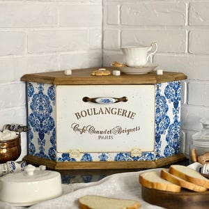 May include: A wooden corner bread bin featuring a white front panel with the text BOULANGERIE Cafe Chaudet Beignets PARIS and a blue and white floral damask pattern on the side panels. The bin has a metal handle and hinges.