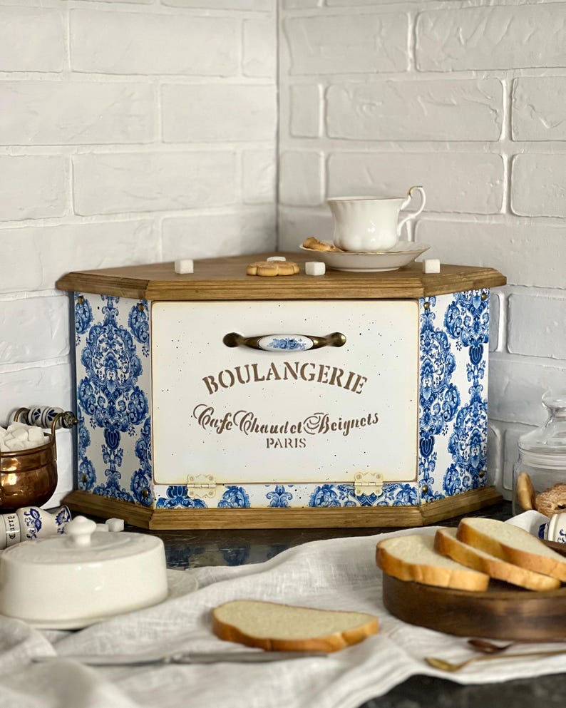 May include: A wooden corner bread bin featuring a white front panel with the text BOULANGERIE Cafe Chaudet Beignets PARIS and a blue and white floral damask pattern on the side panels. The bin has a metal handle and hinges.
