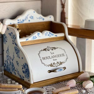 May include: White bread bin with blue floral patterns and the text "La Boulangerie Paris". It features a wooden base and top, a curved lid, and a decorative handle. A cup of tea and biscuits are in the foreground.