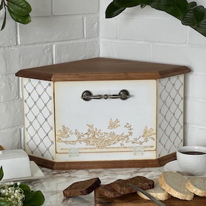 May include: A wooden corner bread bin with a dark brown top and base. The front panel is white with a gold-toned bird and floral illustration, featuring a dark metal handle and two white hinges. The side panels have a grey lattice pattern.