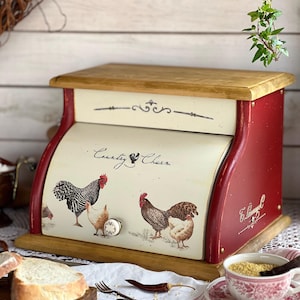 May include: A red and cream-colored bread box with a wooden top and base. The front features a rooster and hen design with the words "Country Charm." The box has a curved, roll-top design and a decorative knob.