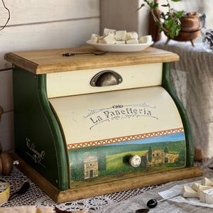 May include: Wooden bread box with a light green and cream color scheme. The box features a curved front with a landscape painting and the text "La Panetteria". A small plate of white cheese sits on top.