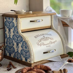 Può includere: Un portapane in legno con uno sportello a serrandina color crema che riporta la scritta BOULANGERIE, Cafe Chaud et Beignets e PARIS. I lati presentano un motivo damascato blu e bianco. Include un cassetto superiore e due maniglie con dettagli floreali.