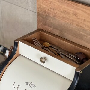 May include: A white and black bread box with a wooden lid and a drawer containing silverware. The box has the words "LE PAIN PARIS" printed on the front. The drawer has a brass knob.