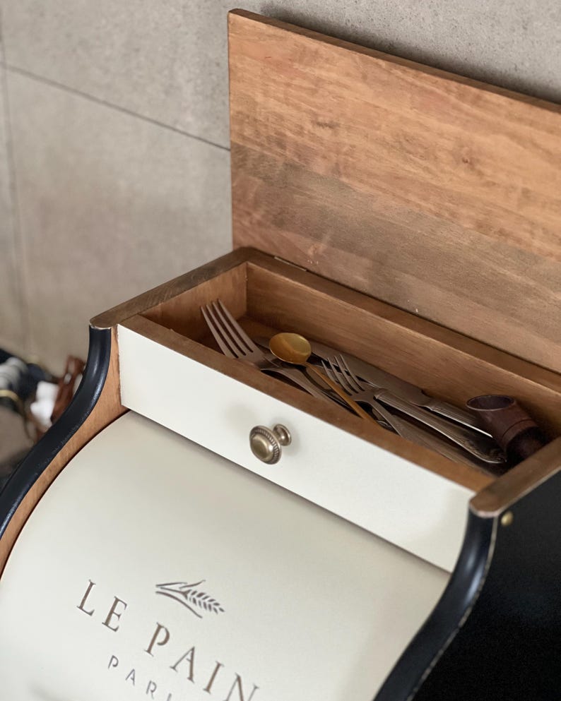 May include: A white and black bread box with a wooden lid and a drawer containing silverware. The box has the words "LE PAIN PARIS" printed on the front. The drawer has a brass knob.