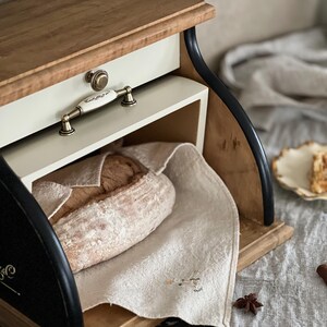 May include: Wooden bread box with a light-colored top and drawer, featuring a cream-colored drawer front and a decorative handle. A loaf of bread rests on a linen cloth inside. The box has black curved side supports.