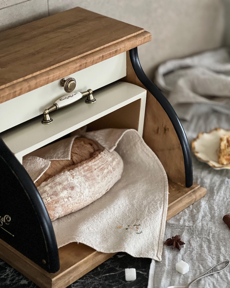 May include: Wooden bread box with a light-colored top and drawer, featuring a cream-colored drawer front and a decorative handle. A loaf of bread rests on a linen cloth inside. The box has black curved side supports.
