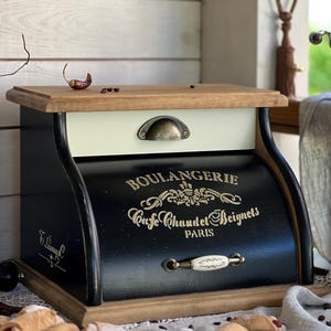 Könnte beinhalten: Ein schwarz-cremefarbener Brotkasten mit Holzdeckel und Boden. Die Vorderseite zeigt den goldenen Schriftzug BOULANGERIE, Cafe Chaud et Beignets und PARIS. Er verfügt über eine cremefarbene Schublade mit Metallgriff und eine schwarze Rolltür mit weißem Griff.
